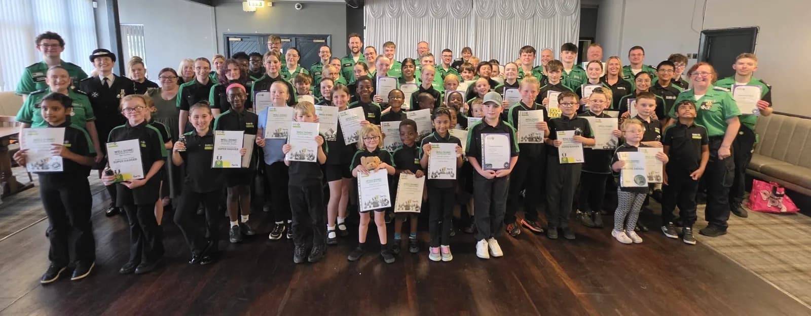 A large group of children and adults, some in uniform, smile and hold up certificates, celebrating an achievement in a brightly lit room.