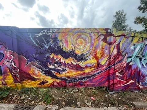 A vibrant, colorful graffiti mural with a swirling yellow and orange vortex at its center, surrounded by purple, black, and red abstract shapes, stretching across a long concrete wall under a cloudy sky.