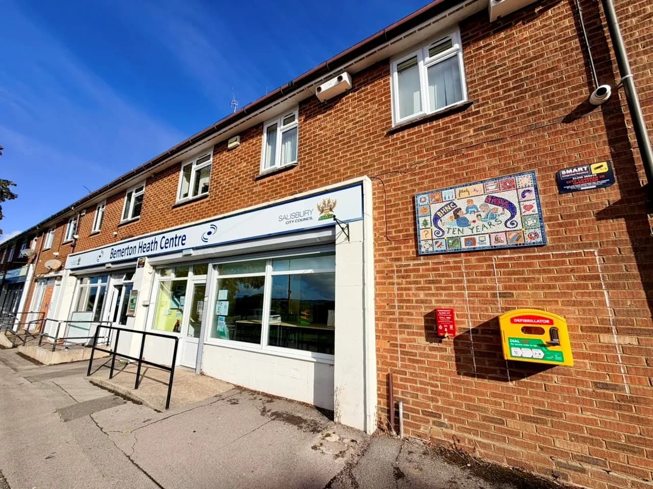 A brick building features a sign for the Bemerton Heath Centre and Salisbury City Council, with a colorful mosaic mural and a defibrillator case mounted on the wall.