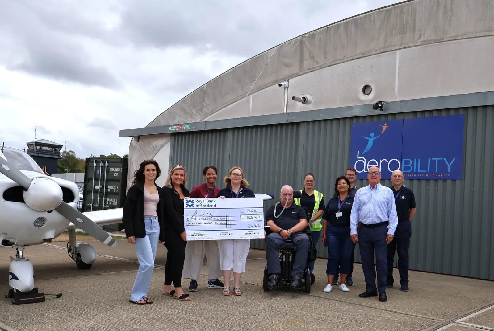 A group of people, some in uniform, stand outside a hangar next to a small airplane, holding a large check.