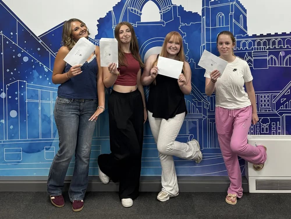 Four young women stand smiling in front of a blue and white mural, each holding an envelope or a piece of paper.