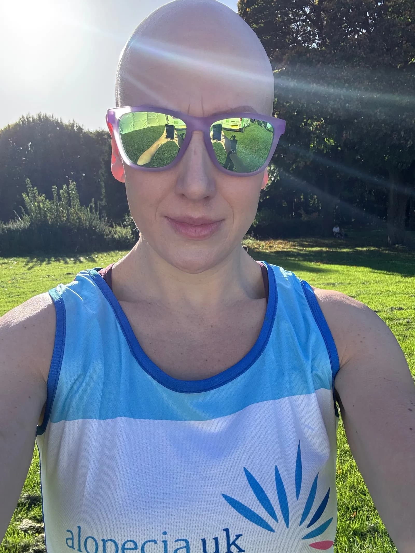 A bald woman in purple sunglasses and an Alopecia UK running vest smiles at the camera with green trees and grass in the background.