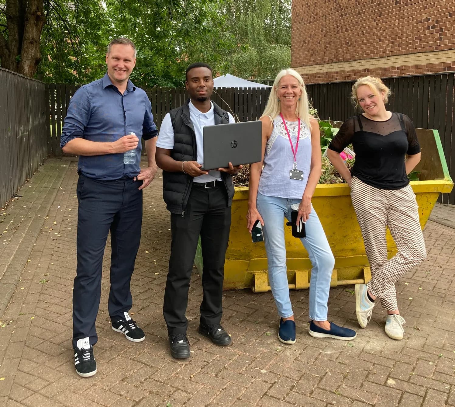 Four people, two men and two women, stand outdoors on a paved area with a yellow skip planter behind them, and one man holds a laptop.