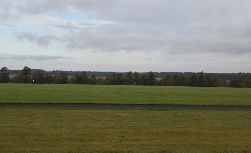 A wide, green field with a dark path running horizontally across the middle, under a cloudy sky.