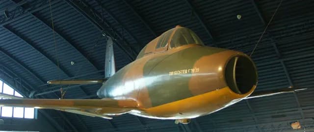 A Gloster Meteor F.Mk.I jet aircraft, painted in camouflage brown, green, and yellow, hangs from a dark ceiling.