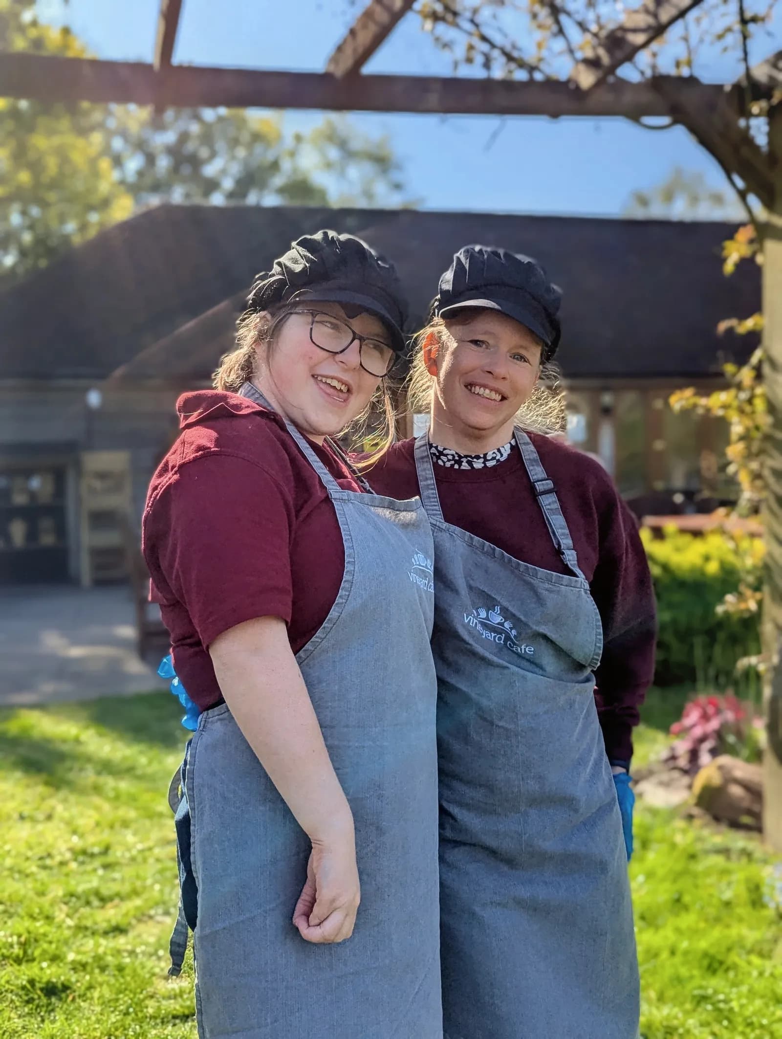 Two women wearing matching dark red shirts, grey aprons, and black caps stand smiling outdoors with a building and greenery in the background.