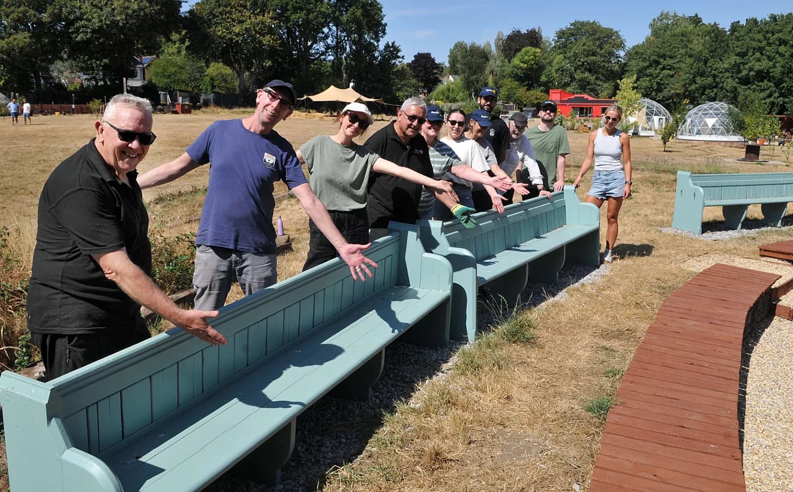 A group of people are standing behind long, light blue benches in a grassy outdoor area, with several smiling and gesturing towards the benches.