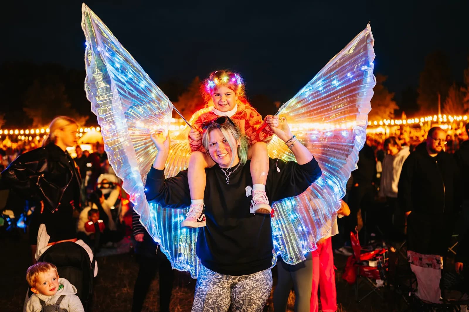 A woman carrying a child on her shoulders, both wearing glowing, colourful outfits, against a blurred background of a night event.
