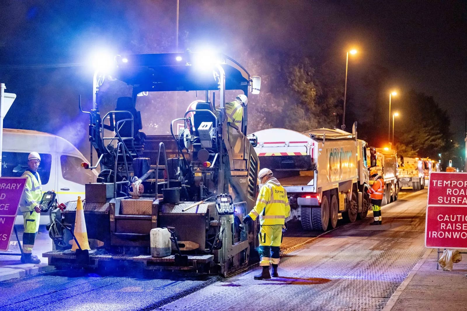 Construction workers in high-visibility gear operate road paving equipment and trucks on a dimly lit street at night.
