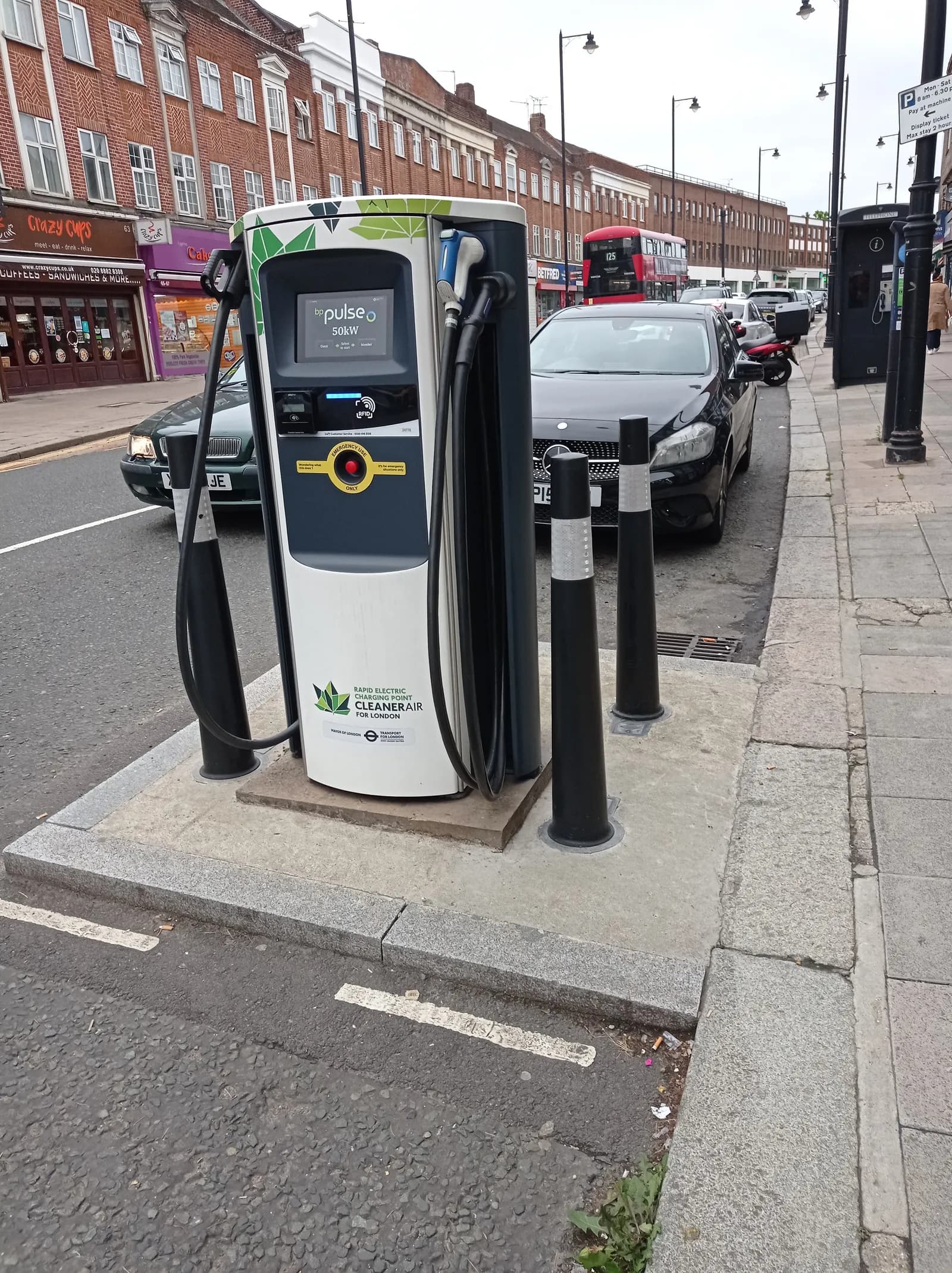 A dual fast electric vehicle charging station stands on a sidewalk, with hoses hanging from its sides, alongside a street with parked cars and storefronts in the background.