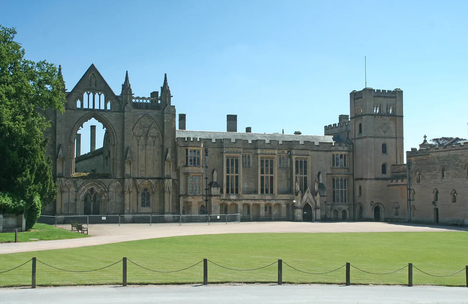 A grand, old stone building with a partially ruined section on the left, an established mansion in the center, and a castle-like tower on the right, all overlooking a wide green lawn.