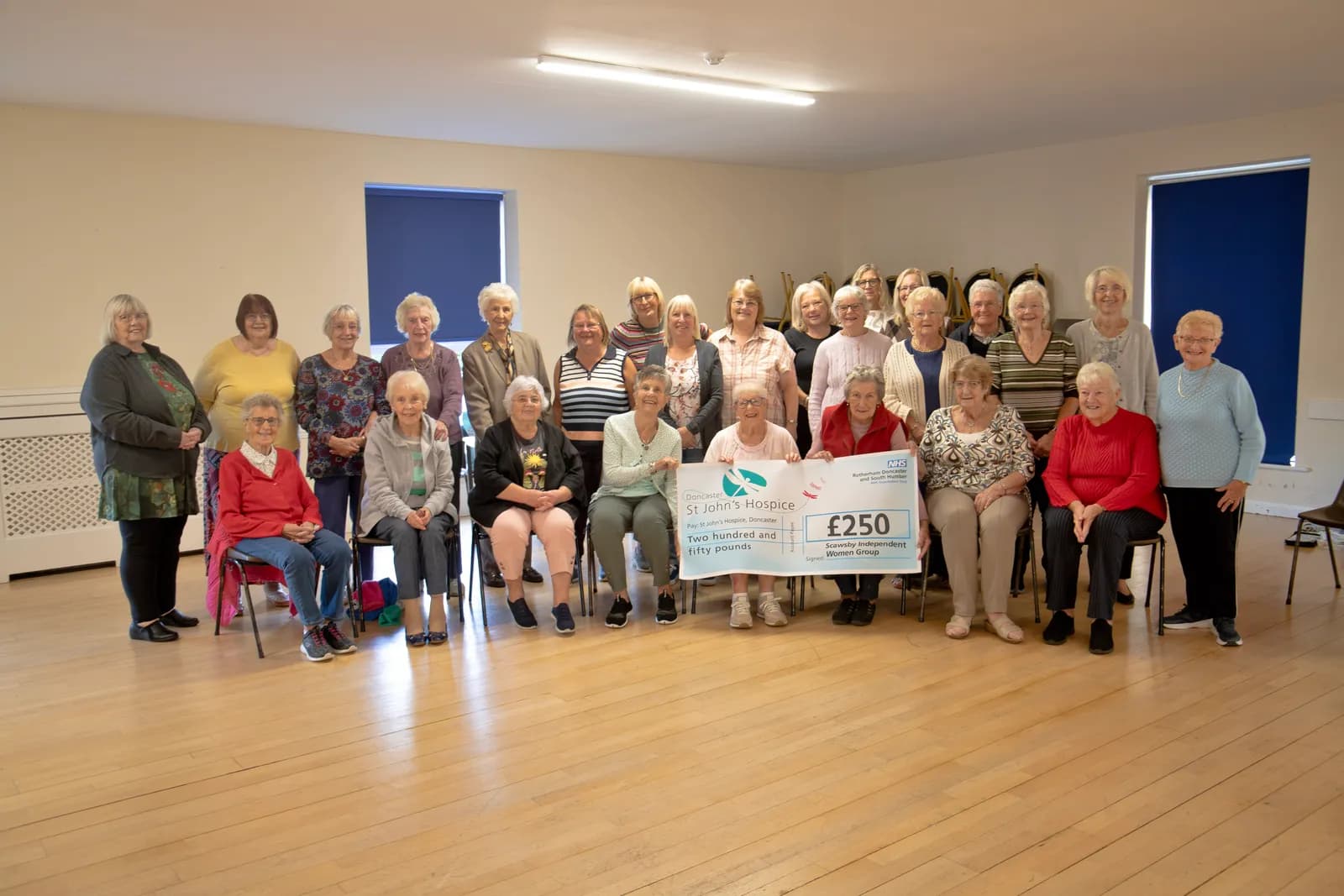 A large group of older women, some sitting and some standing, pose with a large check made out to St John's Hospice for £250.