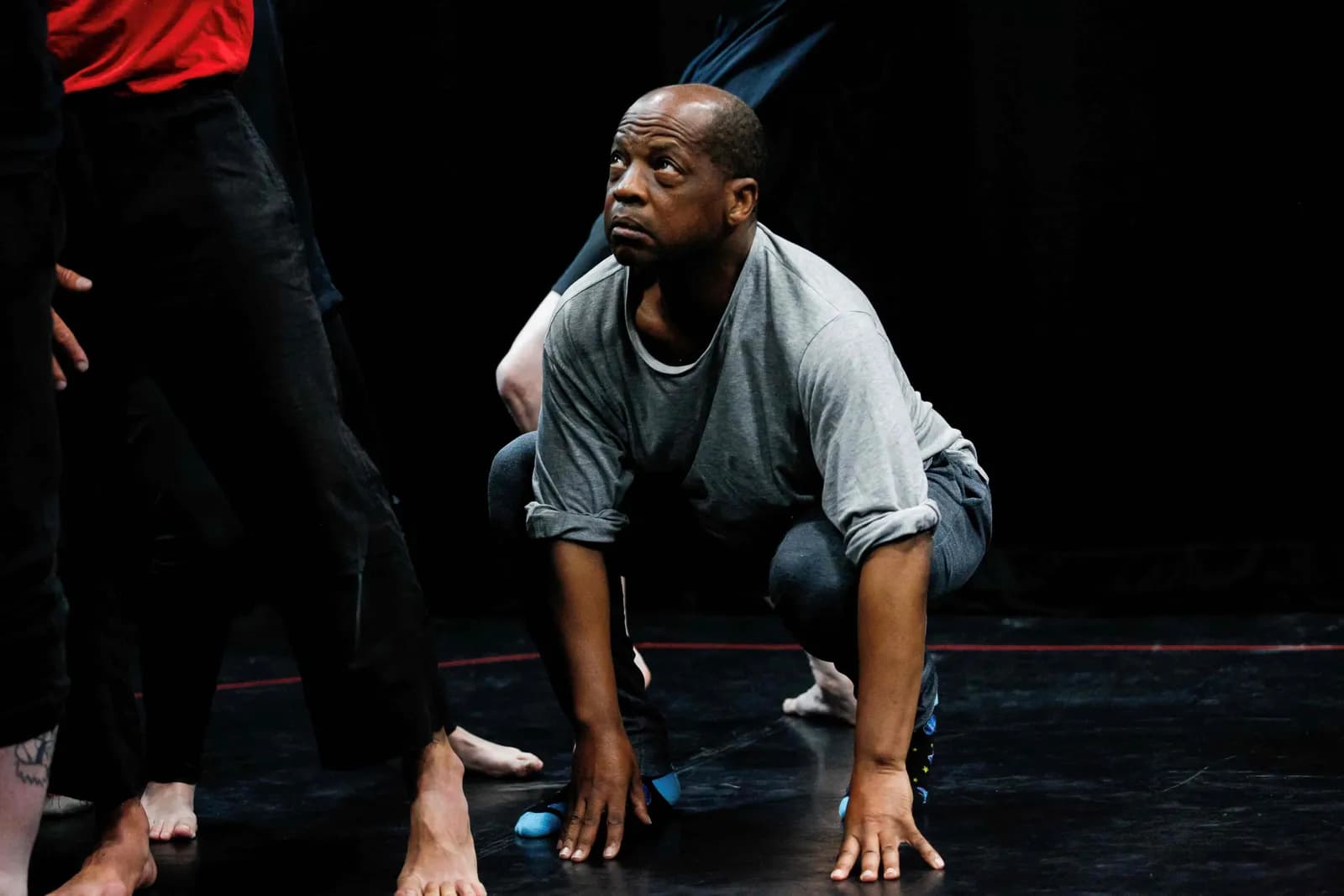 A man in a gray shirt and dark pants crouches on a dark floor, looking up and to the left, surrounded by other people's legs and feet.