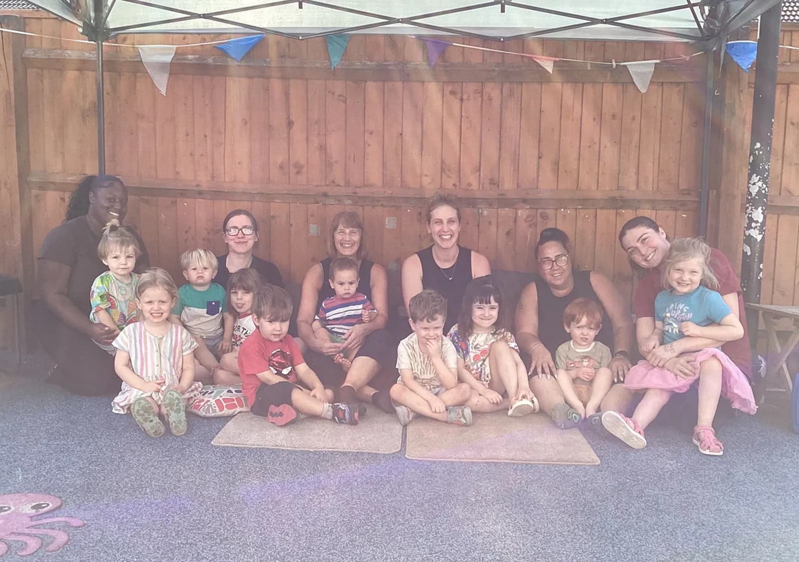 A diverse group of mothers and their young children sit together on the ground, smiling at the camera, with a wooden fence and colorful bunting in the background.