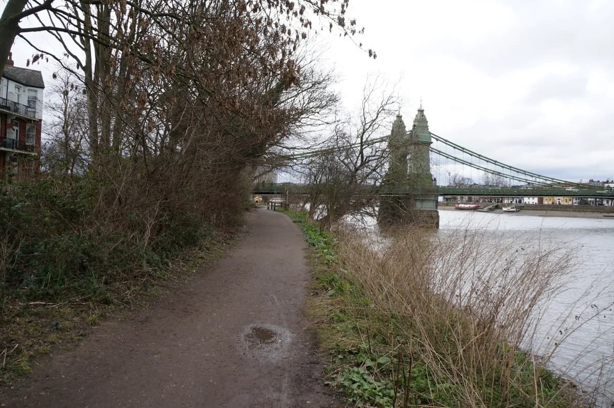 A long, winding dirt path bordered by dense trees and bushes leads towards a large, green suspension bridge crossing a river under a cloudy sky.