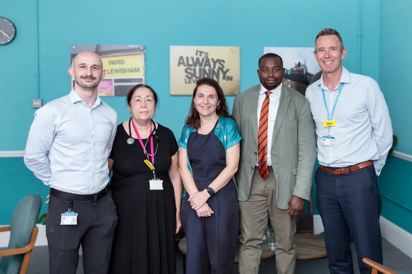 Five people, three men and two women, standing in a line smiling at the camera, with a teal wall and three wall decorations behind them.