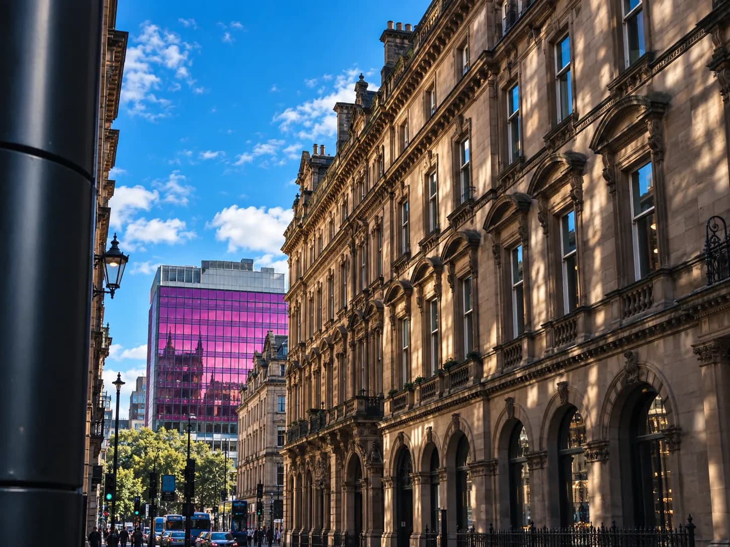A street view showing an ornate, light-colored historic building on the right, and a modern building with a distinct pink glass facade in the distance under a blue sky with white clouds.