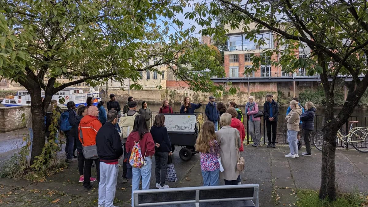 A group of people are gathered outdoors, listening to a woman presenting information on a portable display board next to a river with buildings and boats in the background.