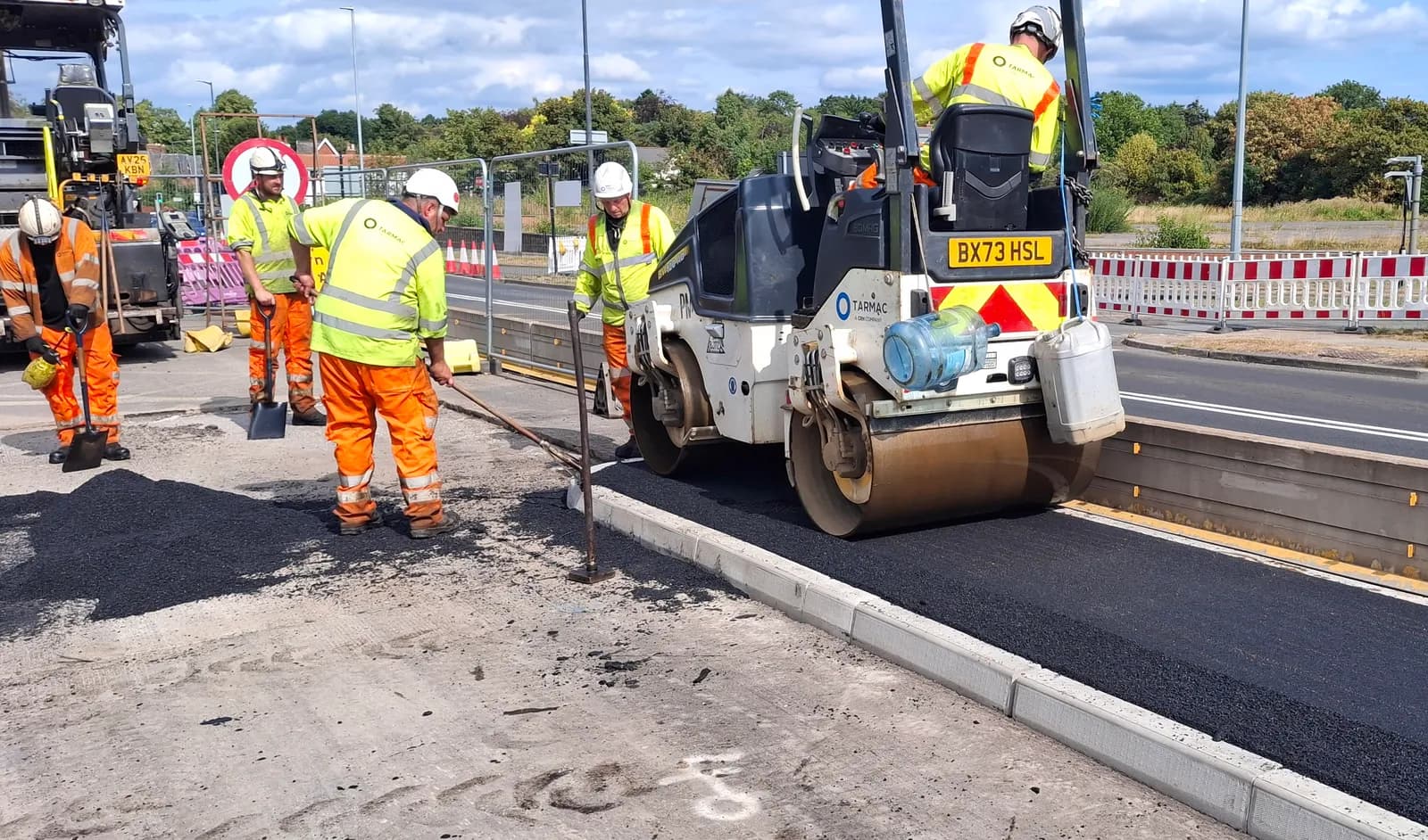 Construction workers in high-visibility clothing and hard hats are paving a road with a steamroller and shovels.