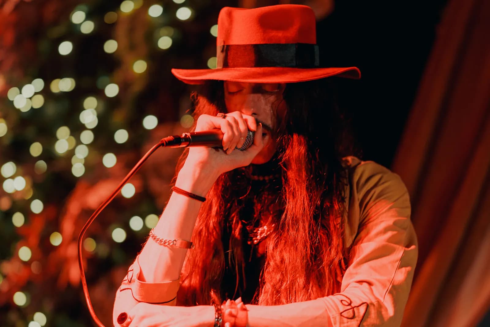 A person with long, dark hair and a red fedora sings into a microphone, illuminated by red and orange lighting with bokeh lights in the background.