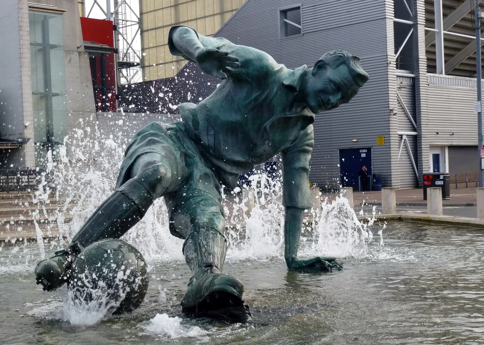 A bronze statue of a soccer player mid-kick in a fountain, with water splashing around the ball and the player's legs, set against a backdrop of modern stadium buildings.