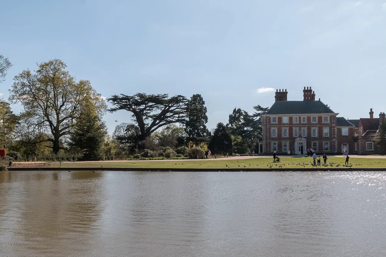 A grand brick estate with multiple chimneys and a white entrance stands partially obscured by trees near a large body of water, with people and geese visible on the grassy bank.