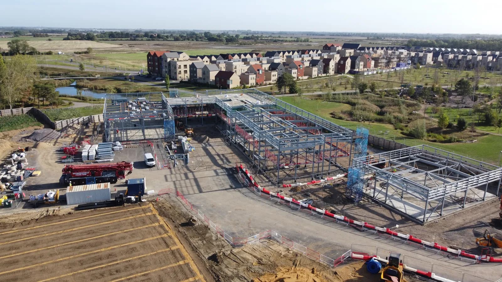 An aerial view of a large construction site with multiple buildings in various stages of steel framework, surrounded by an expanse of green fields and residential homes in the distance.
