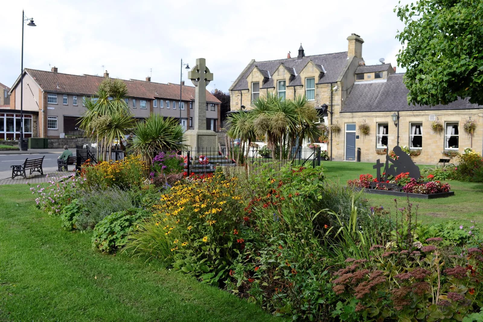 A garden area with colorful flowers and palm trees, a Celtic cross monument, and buildings in the background under a cloudy sky.