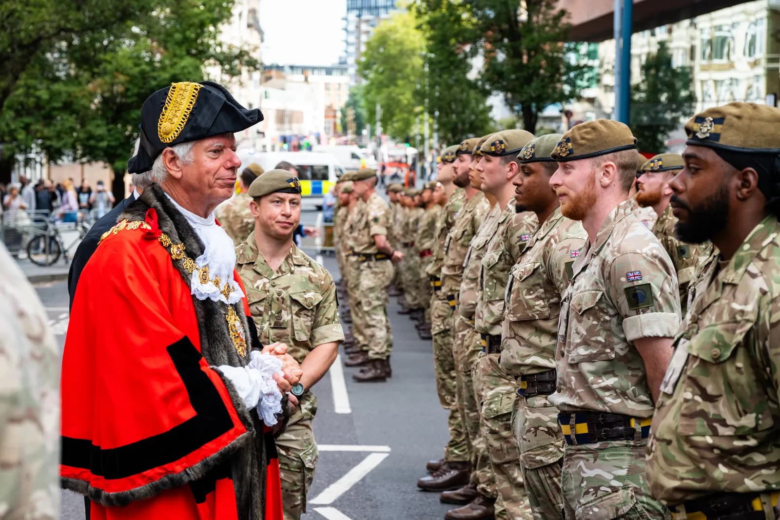 A man in a red and black robe with a bicorne hat and white ruffled cuffs stands in front of a line of soldiers in camouflage uniforms and berets.
