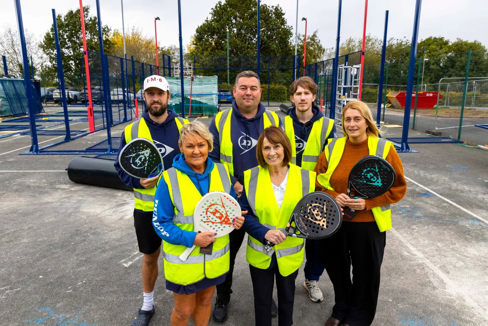 Six people, three men and three women, wearing high-visibility vests and holding padel rackets, stand on a concrete court in front of a partially constructed blue and red court structure.
