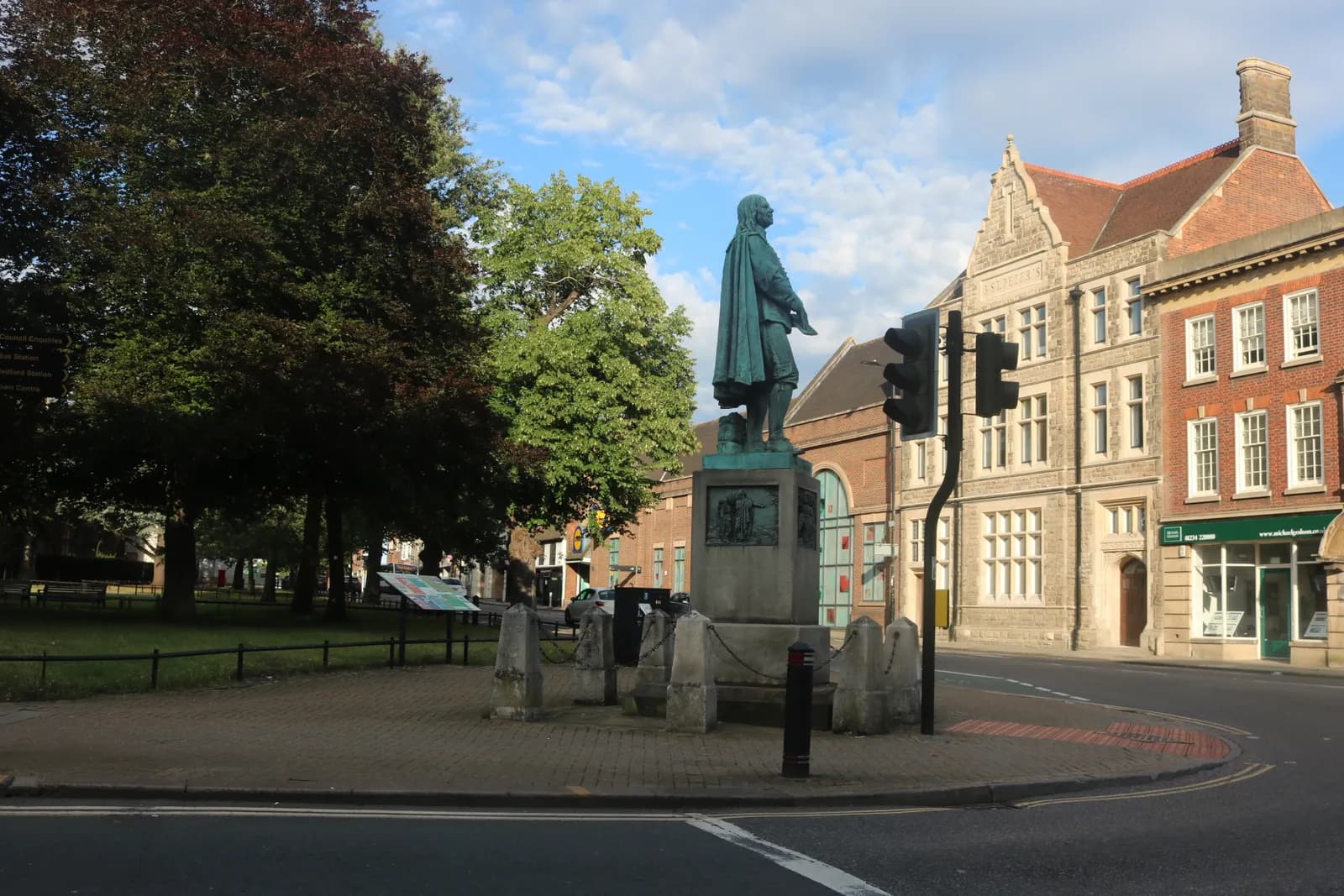 A large bronze statue of a man in historical attire stands in the center of a paved area, flanked by green trees and traditional brick buildings, with a clear sky overhead.