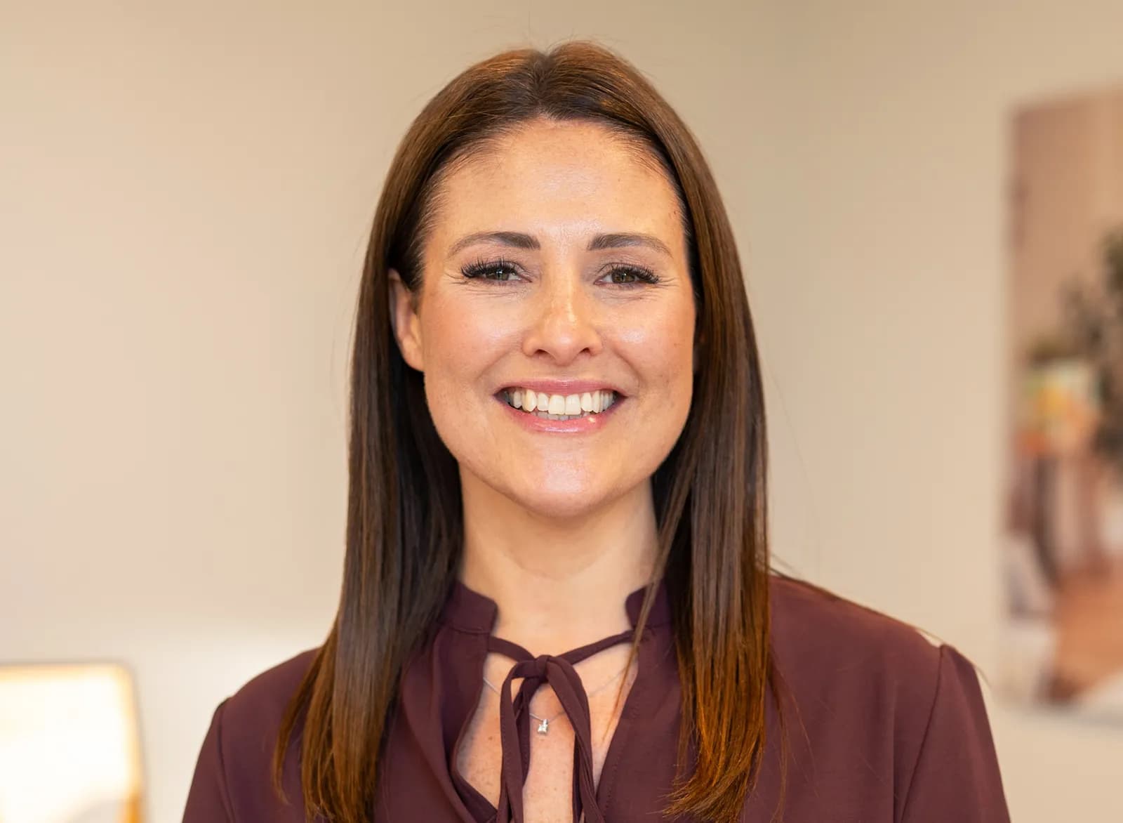 A woman with long brown hair smiles, looking directly at the camera while wearing a dark red collared shirt.