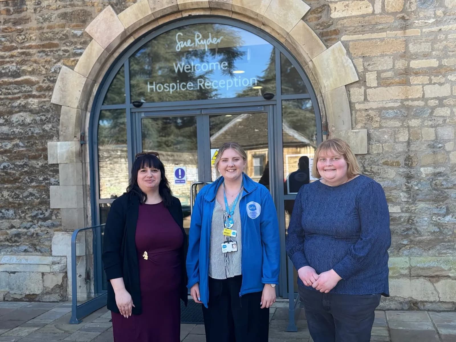 Three women are standing in front of a stone building with an arched glass entrance that reads "Sue Ryder Welcome Hospice Reception."
