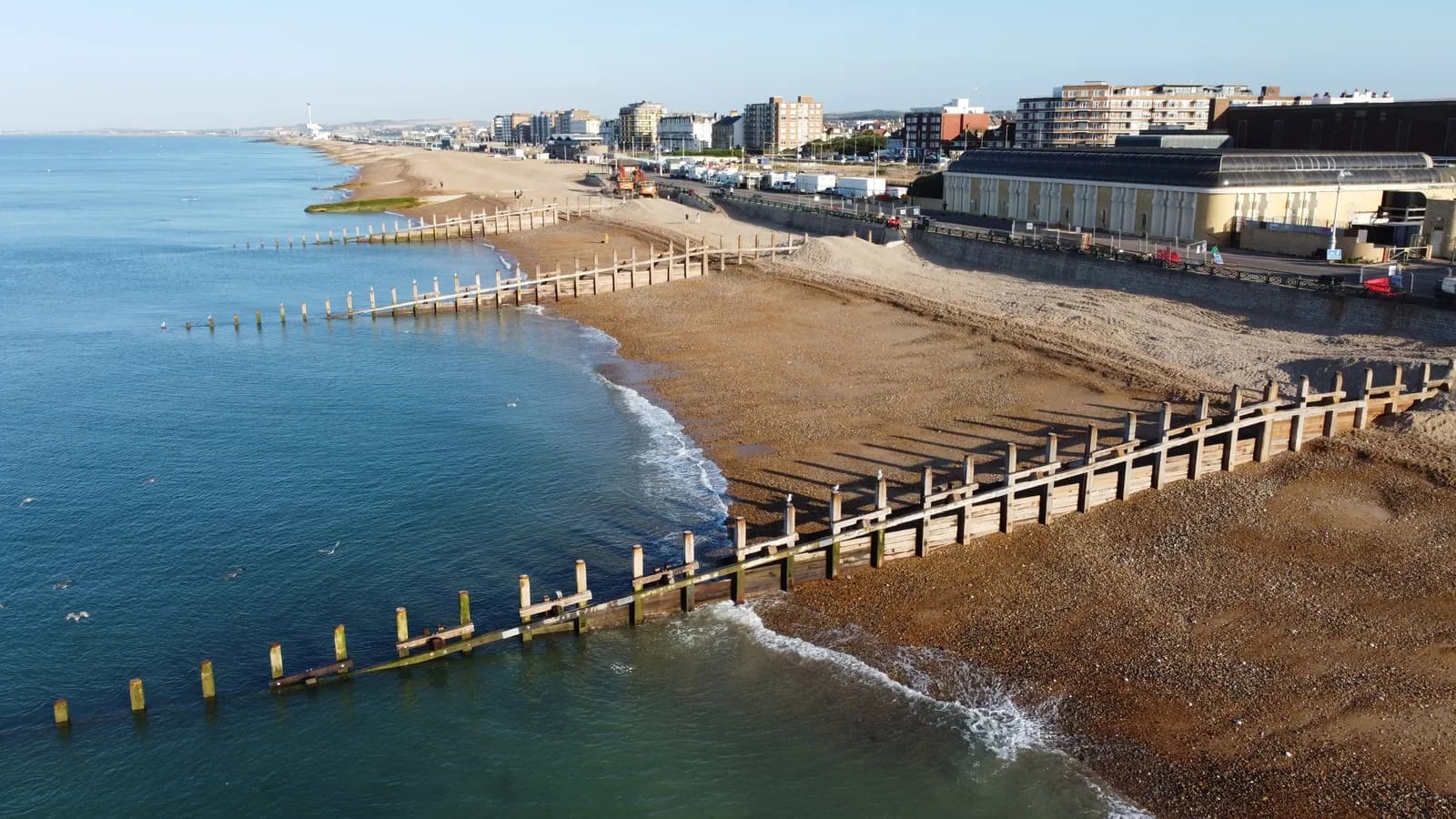 An aerial view of a pebbly beach with wooden groynes extending into the blue ocean, a coastal town with buildings and a promenade visible in the background under a clear sky.