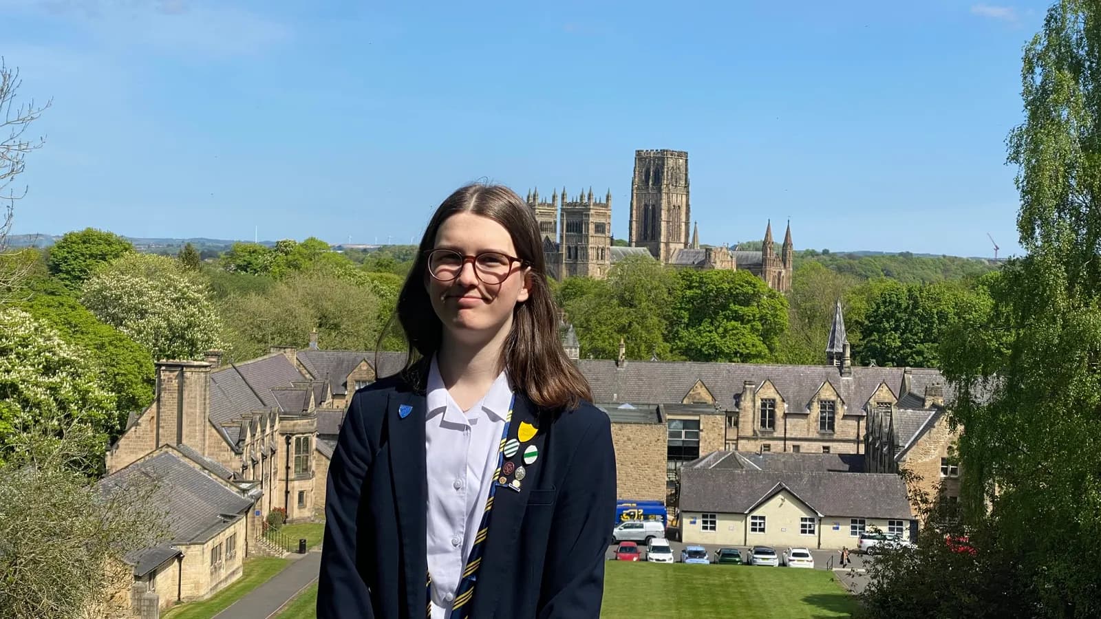 A young woman wearing a school uniform with a blazer and tie stands smiling in front of a distant cathedral and other buildings surrounded by green trees under a clear blue sky.