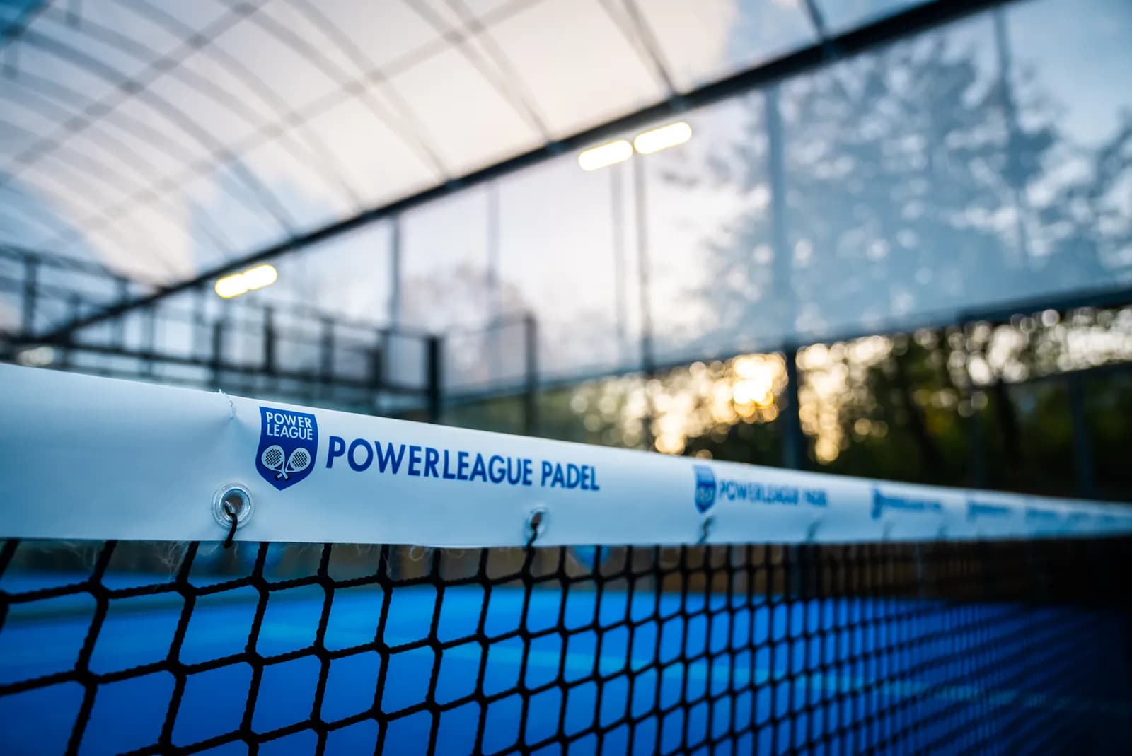 A close-up view of a white banner with the words "POWERLEAGUE PADEL" stretched across a blue and black tennis net, with a blurry background of a court and bright lights.