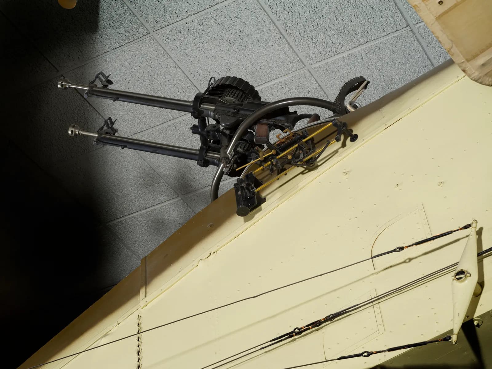 A close-up view of twin black machine guns mounted on the beige-colored wing of an aircraft, with intricate wiring visible against a grey ceiling.