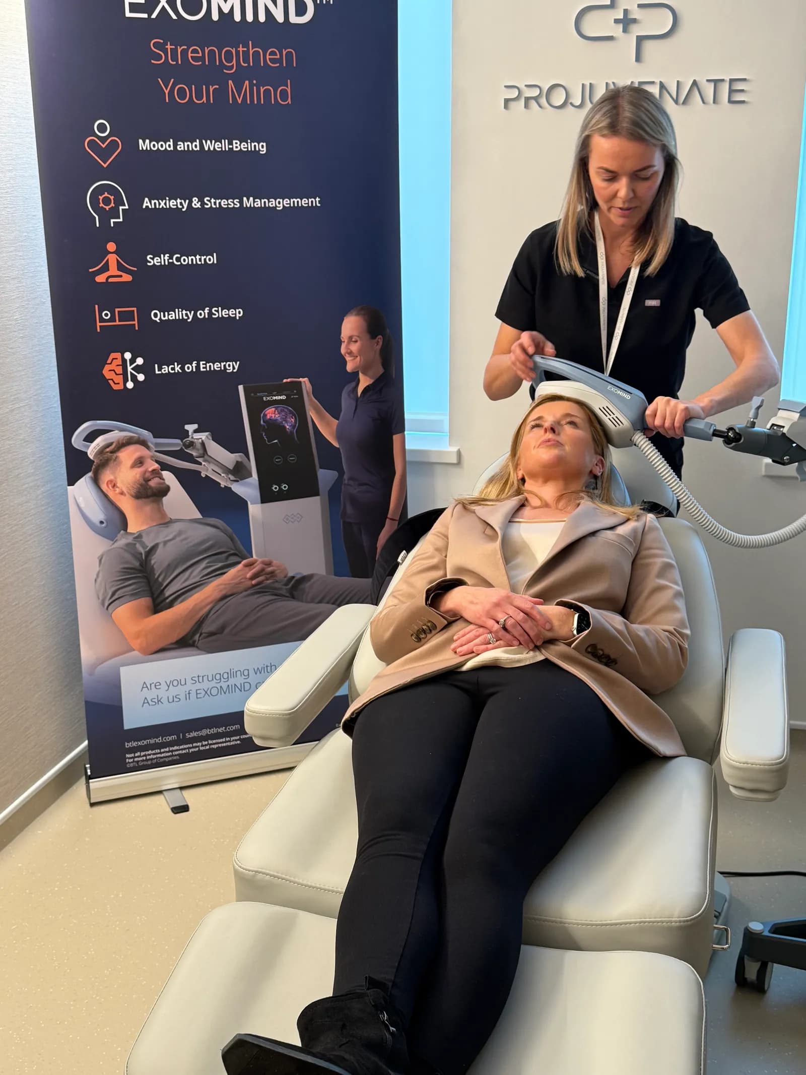 A woman in a black scrub top applies a head device to another woman relaxing in a chair, with a banner for Exomind in the background showing benefits like mood and anxiety management.