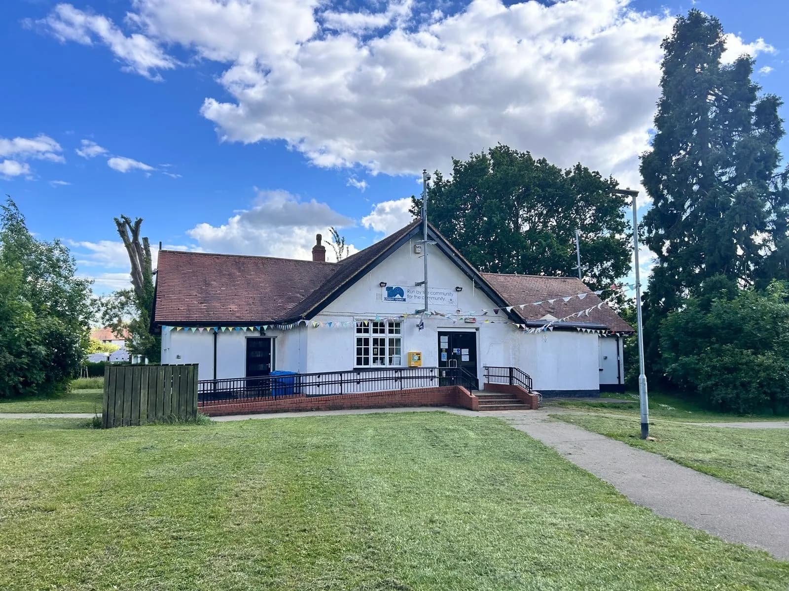A white community building with a brown tiled roof and a ramp leading to the entrance, surrounded by green grass and trees under a blue sky with white clouds.