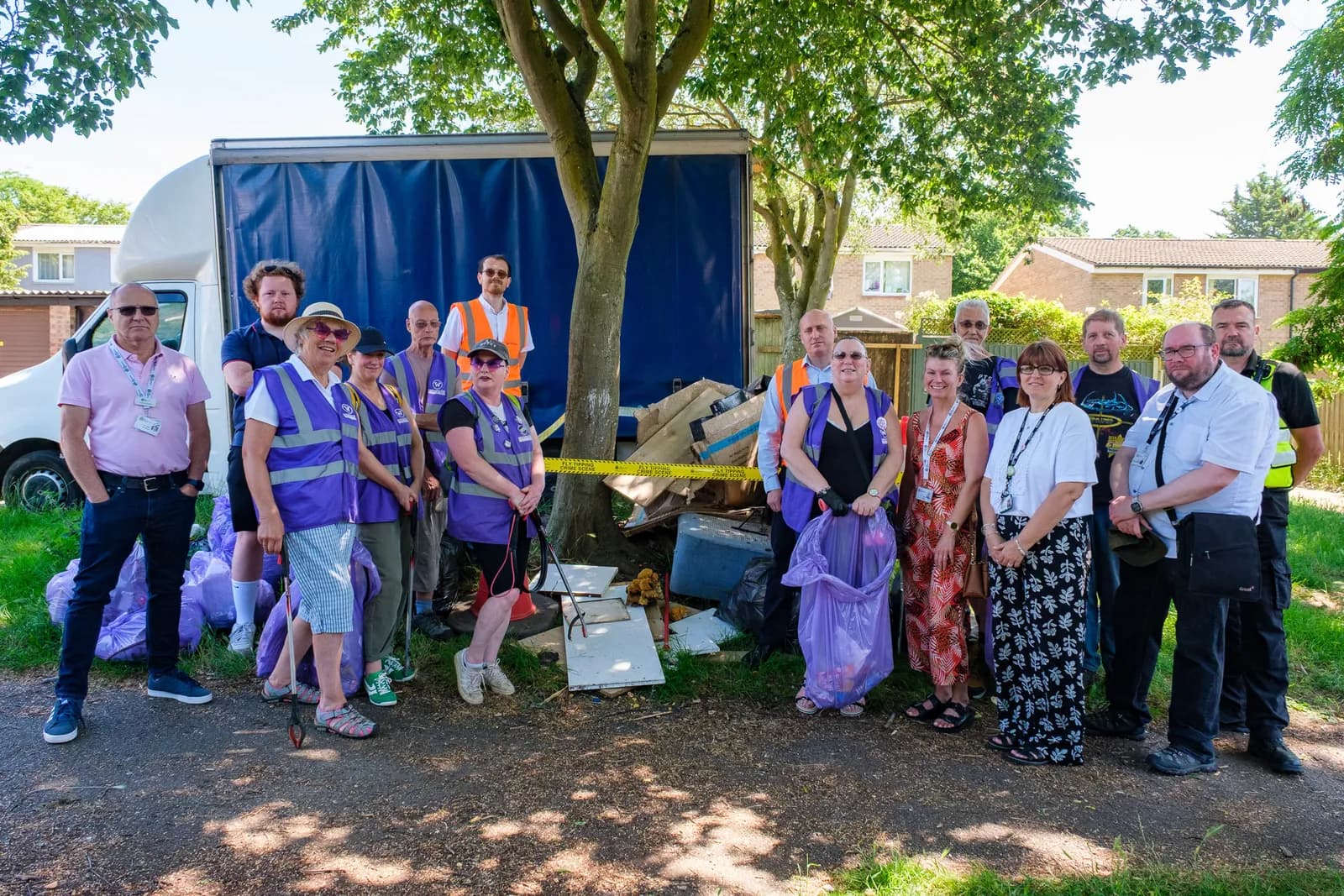 A group of volunteers, some wearing purple vests, stand in front of a blue moving truck and a pile of illegally dumped trash they are cleaning up.