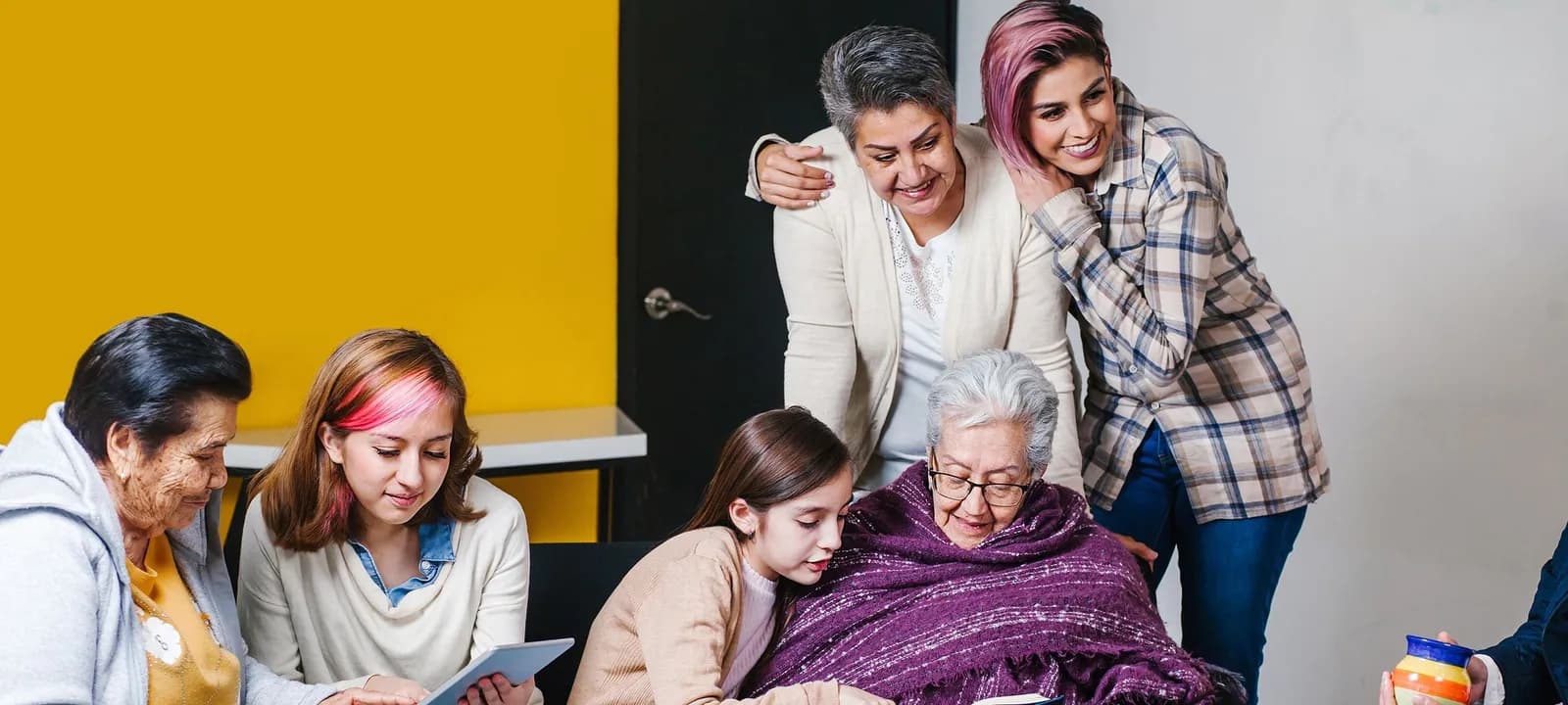 Several generations of women sit around an older woman in a wheelchair, who is covered in a purple blanket; they appear to be reading together.
