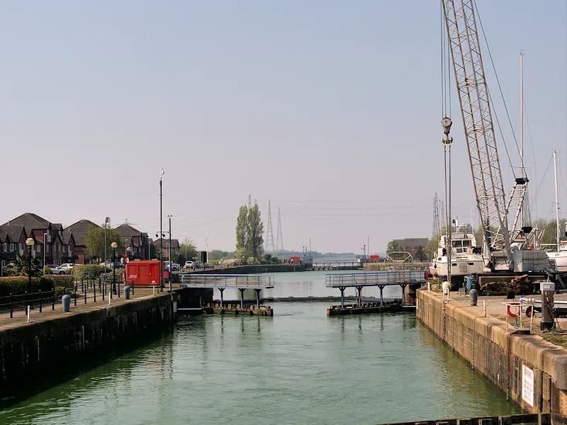 A wide canal with lock gates in the distance, lined by residential buildings on the left and a large industrial crane and boats on the right.
