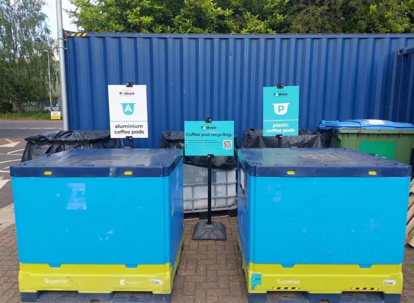Two blue and yellow bins for recycling aluminum and plastic coffee pods are labeled with signs in front of a blue storage container.