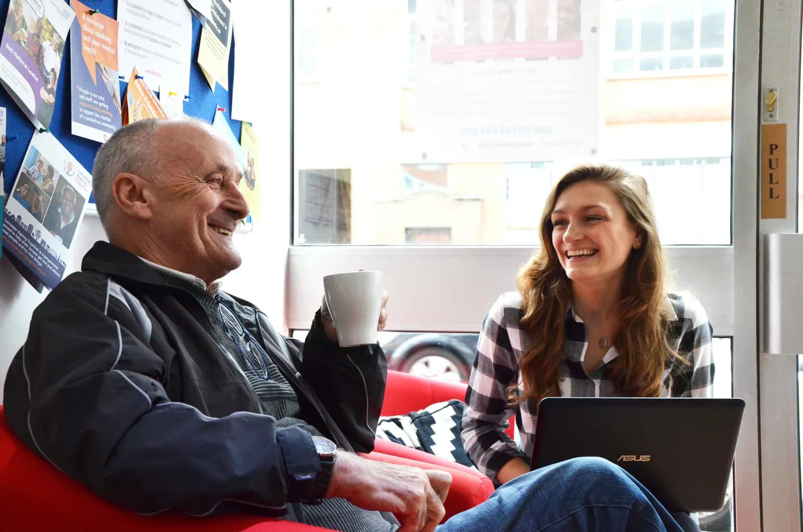 An older man and a younger woman sit together, both smiling and laughing, with the man holding a mug and the woman resting a laptop on her lap.