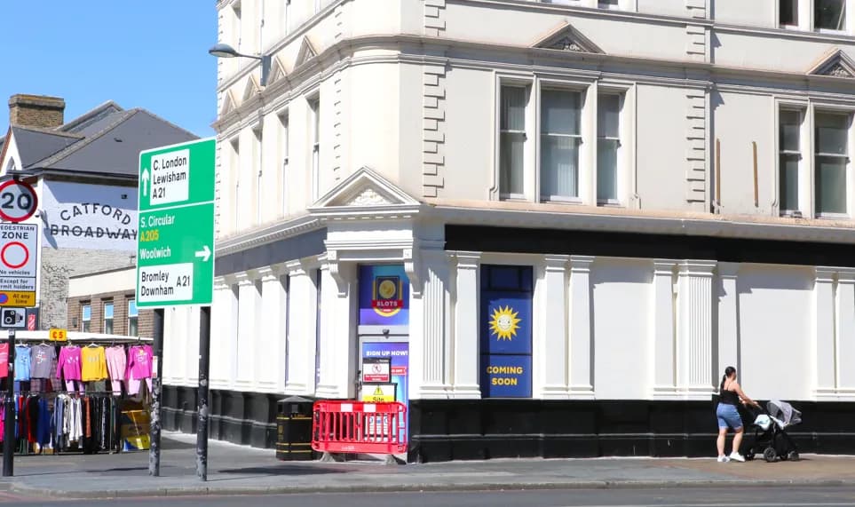 A woman in a black top and blue shorts pushes a baby stroller past a white building with "COMING SOON" and "SLOTS" signs in its windows, with a green street sign for Catford Broadway, Lewisham, and Woolwich visible on the left.