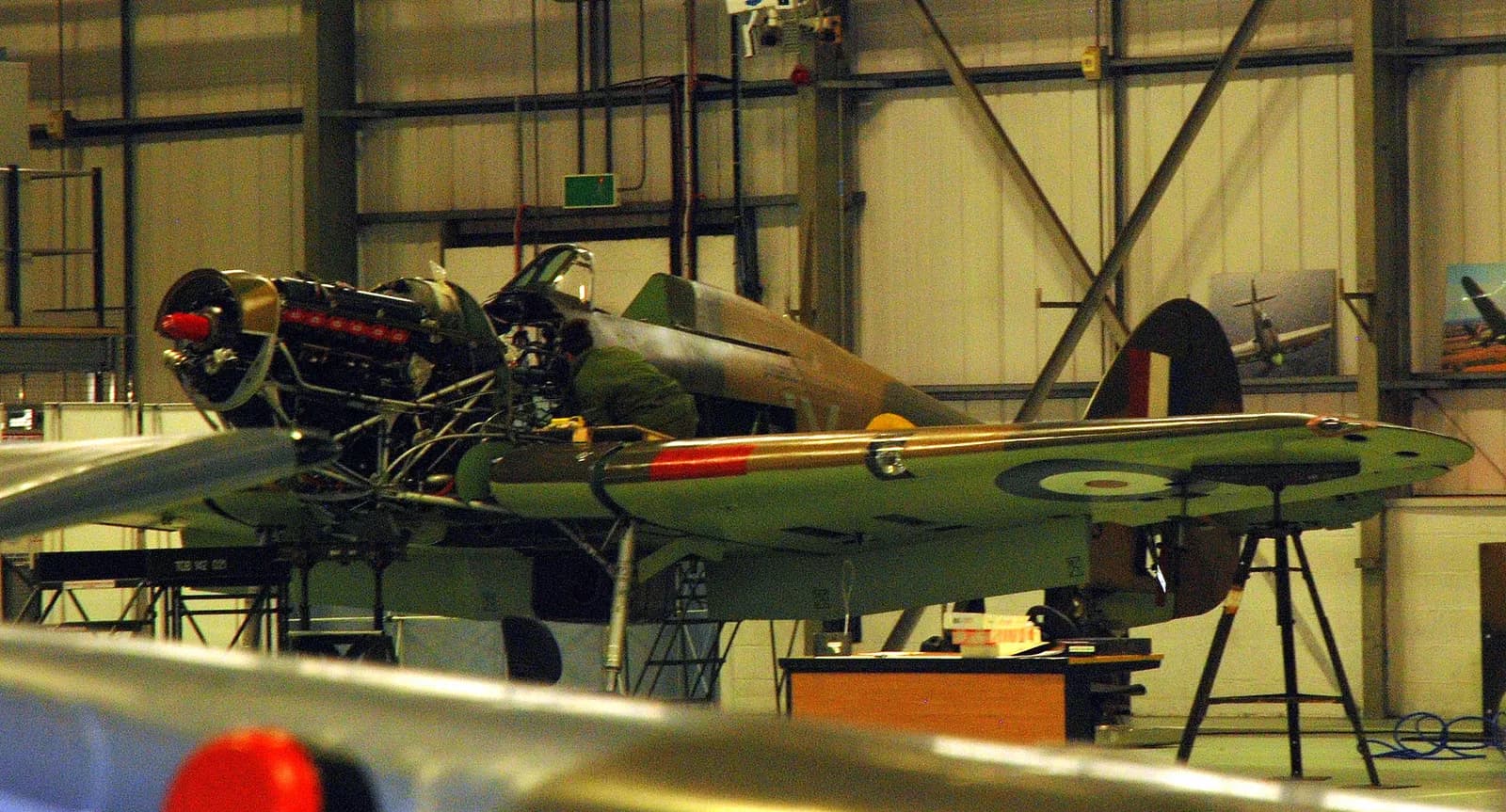 A vintage green military plane is seen from the front, with its engine exposed, on display inside a hangar.
