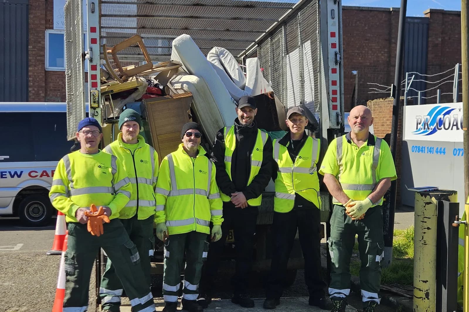 Six men in high-visibility vests and work pants stand smiling in front of a truck loaded with discarded furniture and other items.