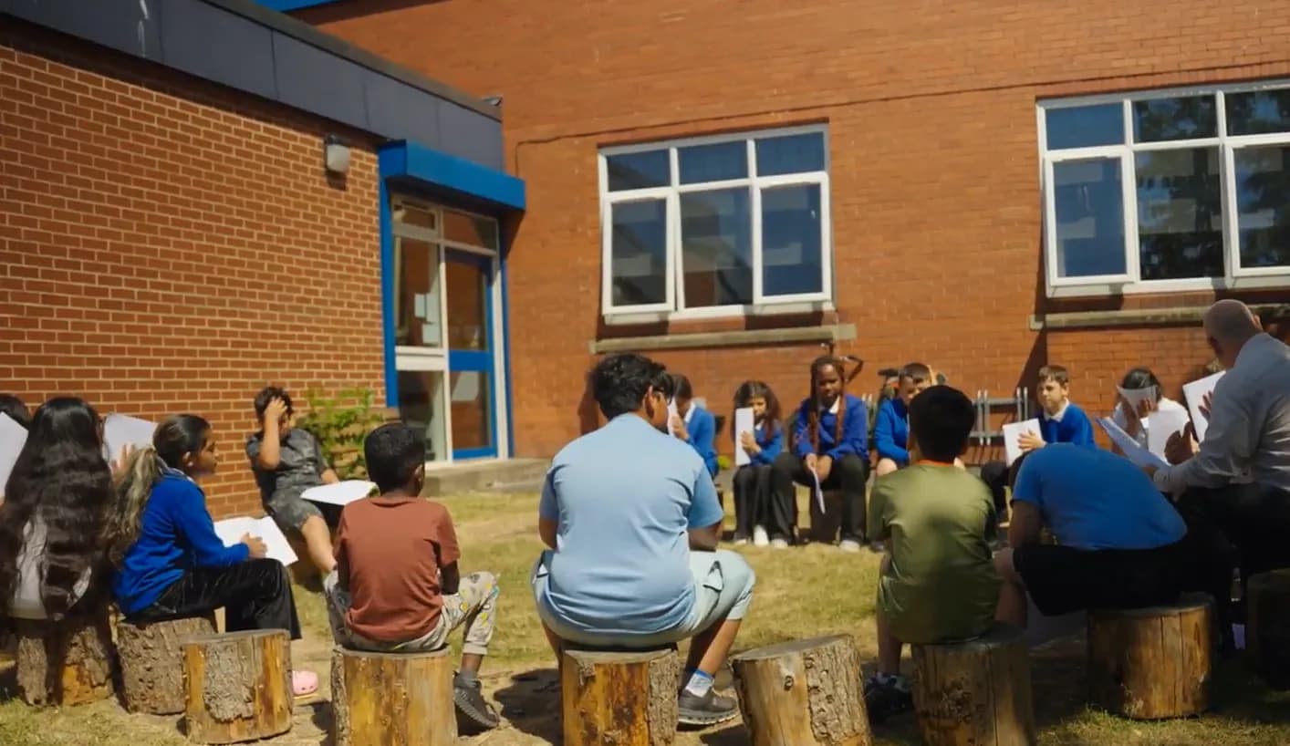 A group of children and adults sit outdoors on wooden stumps in a circle, reading from papers, with a brick building in the background.