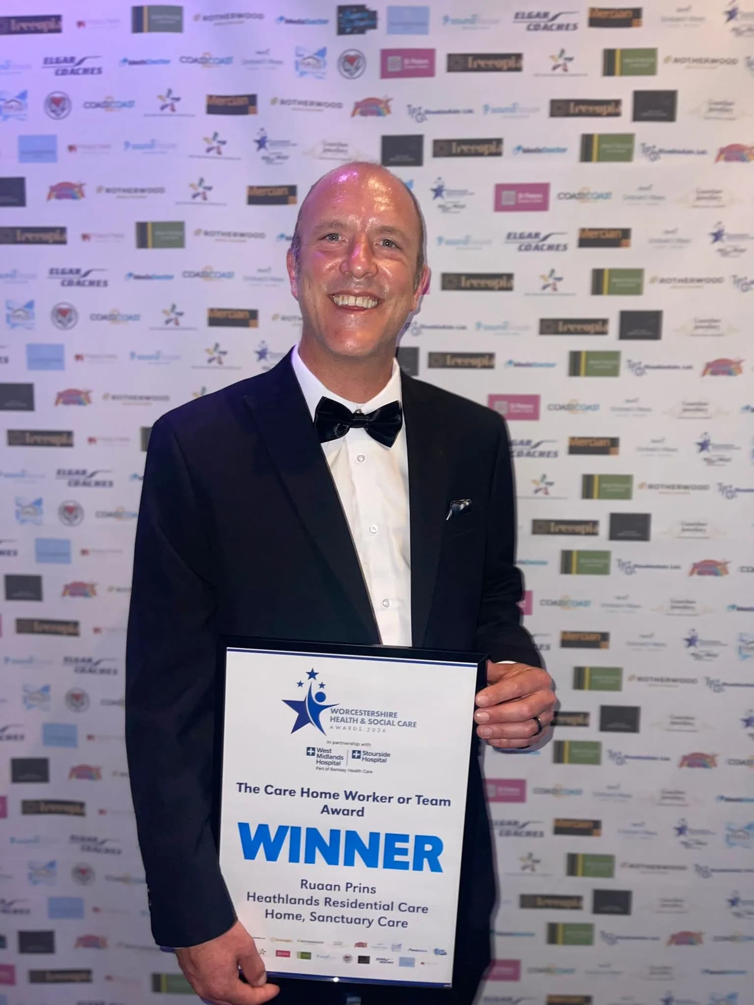 A smiling man in a tuxedo holding a "Winner" award from the Worcestershire Health & Social Care Awards.