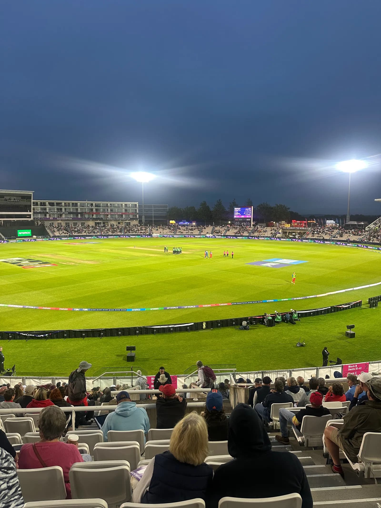 A view from stadium seats of a cricket match being played on a brightly lit green field under a dark gray evening sky.